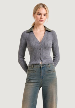 Donna bionda che indossa un cardigan grigio a coste con colletto verde e jeans sbiaditi a vita alta, in piedi con le mani dietro la schiena.