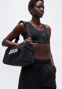 Γυμνασμένο άτομο που φοράει μαύρο αθλητικό σουτιέν και σορτς, κρατώντας μαύρη τσάντα Adidas στον ώμο, στέκεται μπροστά σε απλό φόντο.