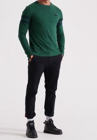 Mann steht mit den Händen in den Taschen, trägt ein grünes Langarmshirt mit dunklen Streifen, schwarze hochgekrempelte Hosen, weiße Socken und schwarze High-Top-Schuhe.