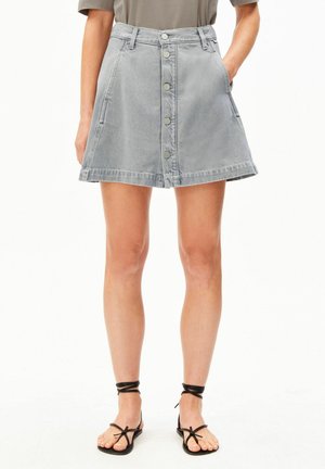 Femme portant une jupe en jean gris clair boutonnée à l'avant avec des poches, associée à des sandales plates noires à brides et un haut vert olive rentré.