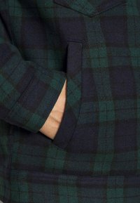 Zielono-granatowa kurtka w kratę, z przednią kieszenią, teksturowanym materiałem i klasycznym kołnierzem. Rękawy są ciemne i starannie wykończone.