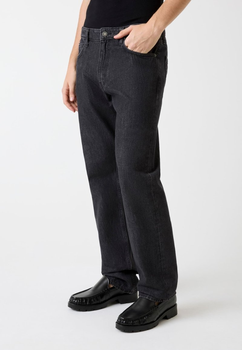 Jean en denim noir à coupe décontractée, conception à cinq poches et fermeture par bouton, associé à des mocassins noirs. Texture lisse, détails minimaux.