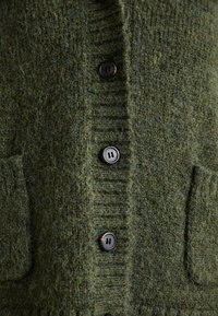 Overgroot groen gebreid vest met gestructureerde stof, ribbels aan de randen, twee voorzakken en zwarte knopen.