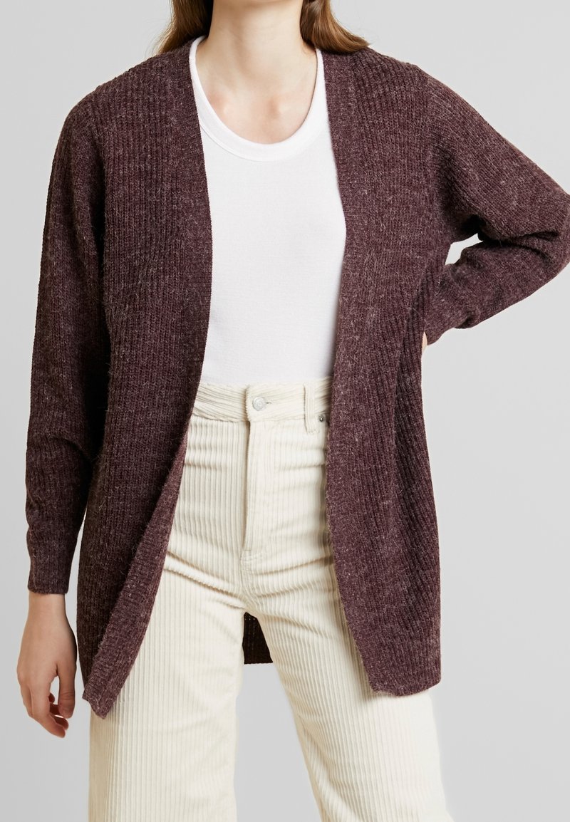 Viininpunainen villatakki, jossa on ribbikuviointi, auki oleva malli ja pitkät hihat. Käytetään valkoisen paidan päällä, yhdistettynä beigeihin corduroy-housuihin.