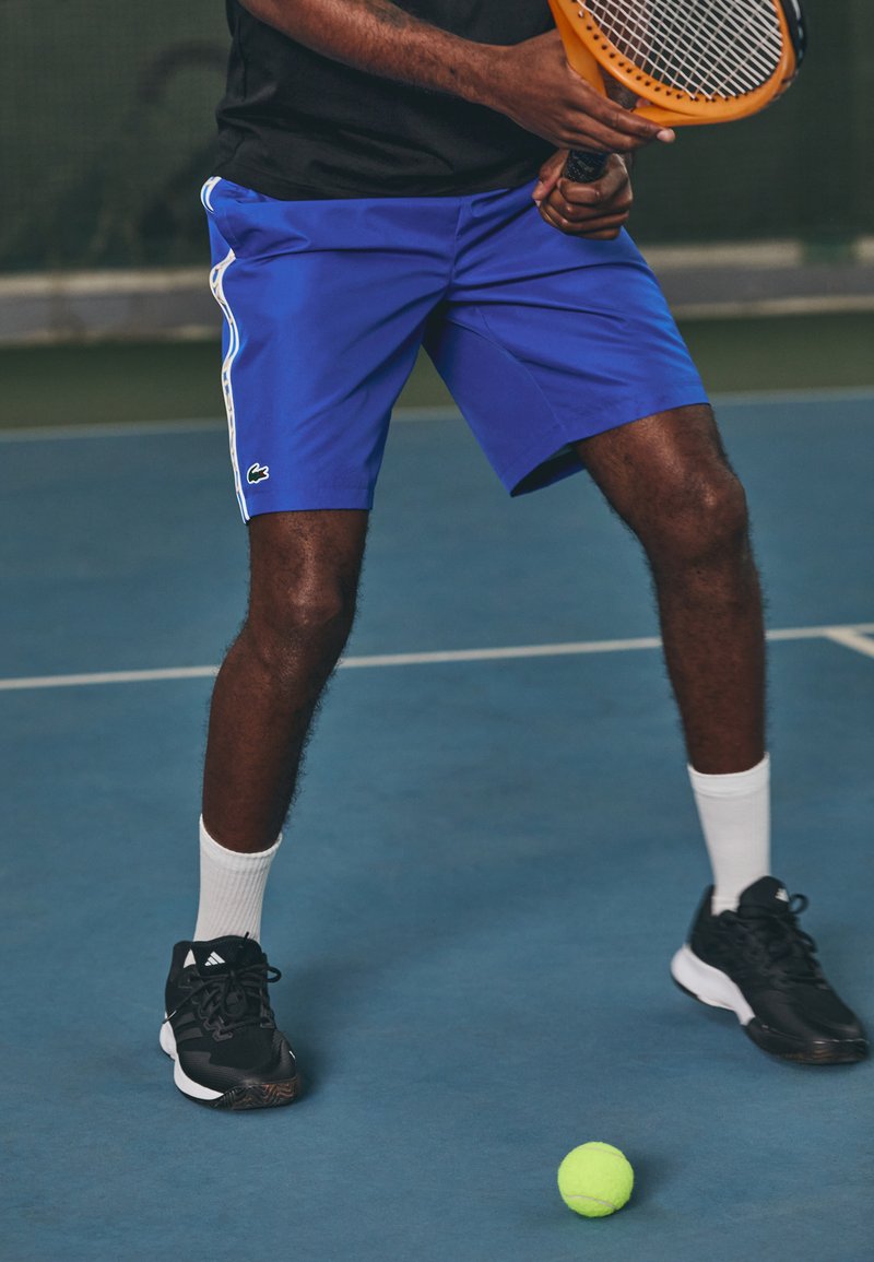 Μπλε αθλητικά σορτς με άσπρες πλευρικές ρίγες, μαύρα αθλητικά παπούτσια, άσπρες κάλτσες και ένα ρακέτα τένις; μια πράσινη μπάλα τένις στο γήπεδο.