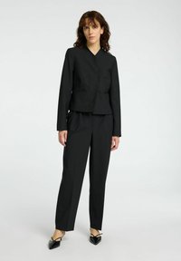 Černý šitý blazer s výstřihem do V, dvěma předními kapsami a odpovídajícími kalhotami. Materiál se zdá být hladký s strukturovaným designem. Špičaté černé boty.