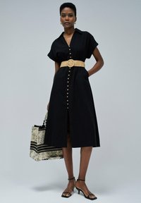 Schwarzes, kurzärmliges Hemdblusenkleid, tailliert mit einem geflochtenen Gürtel. Verfügt über einen Schlitz vorne. Begleitet von einer gemusterten Tragetasche.