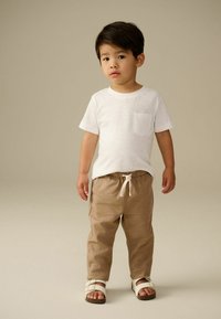 T-shirt bianco in cotone con taschino sul petto, abbinato a pantaloni di lino marrone chiaro con cintura elastica, completato da sandali bianchi.