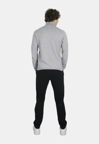 Uomo con capelli ricci che indossa un maglione grigio chiaro a collo alto, pantaloni neri e sneakers bianche, in piedi con le mani in tasca e rivolto di spalle.