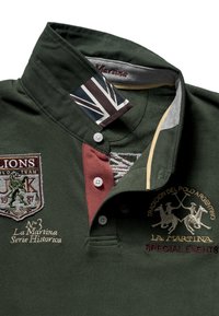 Tumma vihreä poolopaita, jossa on teksturoitu kangas, viininpunainen nappilista, kirjailtut logot ja Union Jack -koriste kauluksessa.