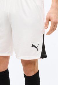 Λευκά αθλητικά σορτς με μαύρες πλαϊνές λεπτομέρειες και λογότυπο Puma, φορεμένα με μαύρες κάλτσες μέχρι το γόνατο, εμφανίζοντας τα πόδια και το δεξί χέρι.
