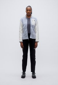 Ανοιχτό μπλε και κρεμ φλις μπουφάν με τσεπάκι με φερμουάρ, συνδυασμένο με μαύρο παντελόνι και γκρι παπούτσια, εμφανίζεται με πρόσωπο προς τα εμπρός σε απλό φόντο.