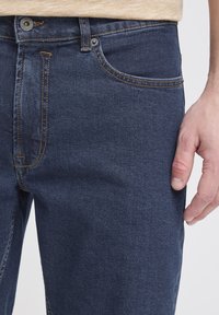 Tumma siniset denim-farkut, joissa klassinen viiden taskun malli, kontrastivärisillä oransseilla tikkauksilla ja vetoketjulla varustettu etuosa, jossa metallinen nappi.