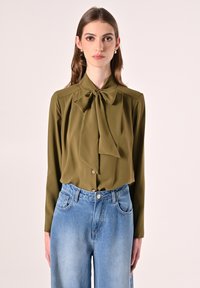 Olivegrüne Bluse mit einem Schleifenkragen, langen Ärmeln und Knopfleiste; kombiniert mit hoch taillierten hellblauen Jeans.