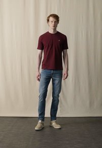 Burgundi rövid ujjú póló, világoskék farmernadrág és bézs sportcipő. A pólón egy kisebb logó található; az öltözet lazán illeszkedik.