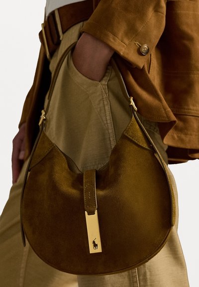 Smeđa nubuk torba za rame s zakrivljenim oblikom, s zlatnim metalnim naglaskom i oznakom s logotipom. Glatka tekstura i jedna ručka.