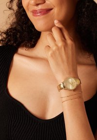 Goldene Uhr mit rundem Ziffernblatt, Kristallakzenten und einem metallischen Band. Trägt sie an einem Handgelenk mit einem zarten goldenen Kettenarmband. Strukturierter schwarzer Oberteil.