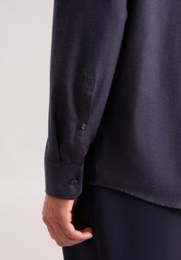 Maglietta a maniche lunghe blu navy con polsini abbottonati e tessuto testurizzato. Include un piccolo logo vicino al polsino. Finitura liscia e vestibilità su misura.