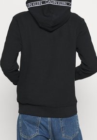 Felpa nera con cappuccio in misto cotone. Presenta un band e logo bianco sul cappuccio e una vestibilità rilassata, abbinata a jeans di denim blu.