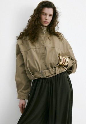 Mujer con pelo largo y rizado que lleva una chaqueta beige con cinturón, pantalones negros de pierna ancha y grandes brazaletes dorados en su brazo izquierdo.