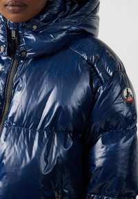 Giacca puffer blu navy con finitura lucida, caratterizzata da un colletto alto, chiusura con zip e tasche laterali. Logo del marchio sulla manica sinistra.