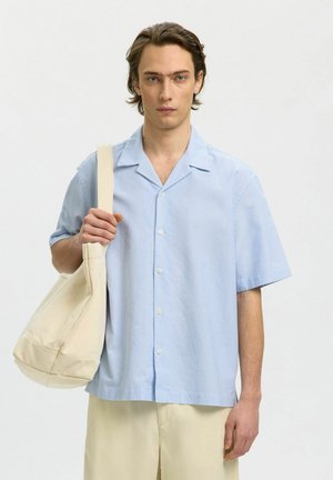 Ung mand iført en lyseblå kortærmet skjorte med knapper og beige bukser, der bærer en stor beige tote-taske over den ene skulder.