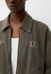 Khaki džínová bunda se zipem vpředu, špičatým límcem a vyšitými logy. Tkanina má mírně strukturovaný povrch.