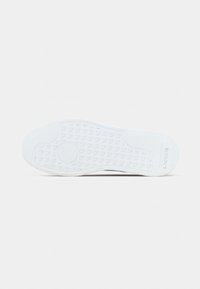 Λευκή σόλα παπουτσιού με υφή λαβής και το λογότυπο "LACOSTE" στην περιοχή της φτέρνας σε απλό φόντο.