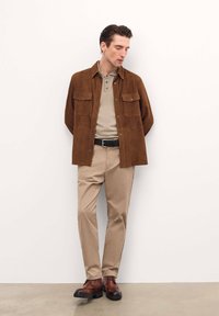 Giacca camicia in suede marrone con due tasche sul petto, polo beige, pantaloni beige e scarpe in pelle marrone con suola nera.