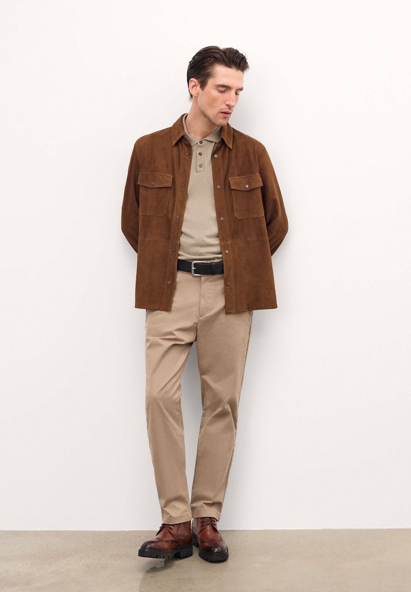 Giacca camicia in suede marrone con due tasche sul petto, polo beige, pantaloni beige e scarpe in pelle marrone con suola nera.