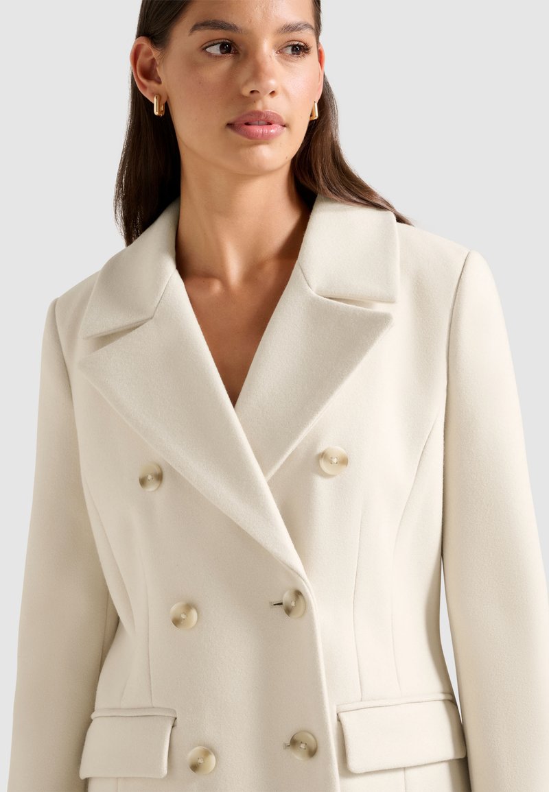 Cappotto bianco doppiopetto realizzato in tessuto morbido con grandi bottoni color crema, rever sharp e tasche laterali, che presenta una silhouette aderente.