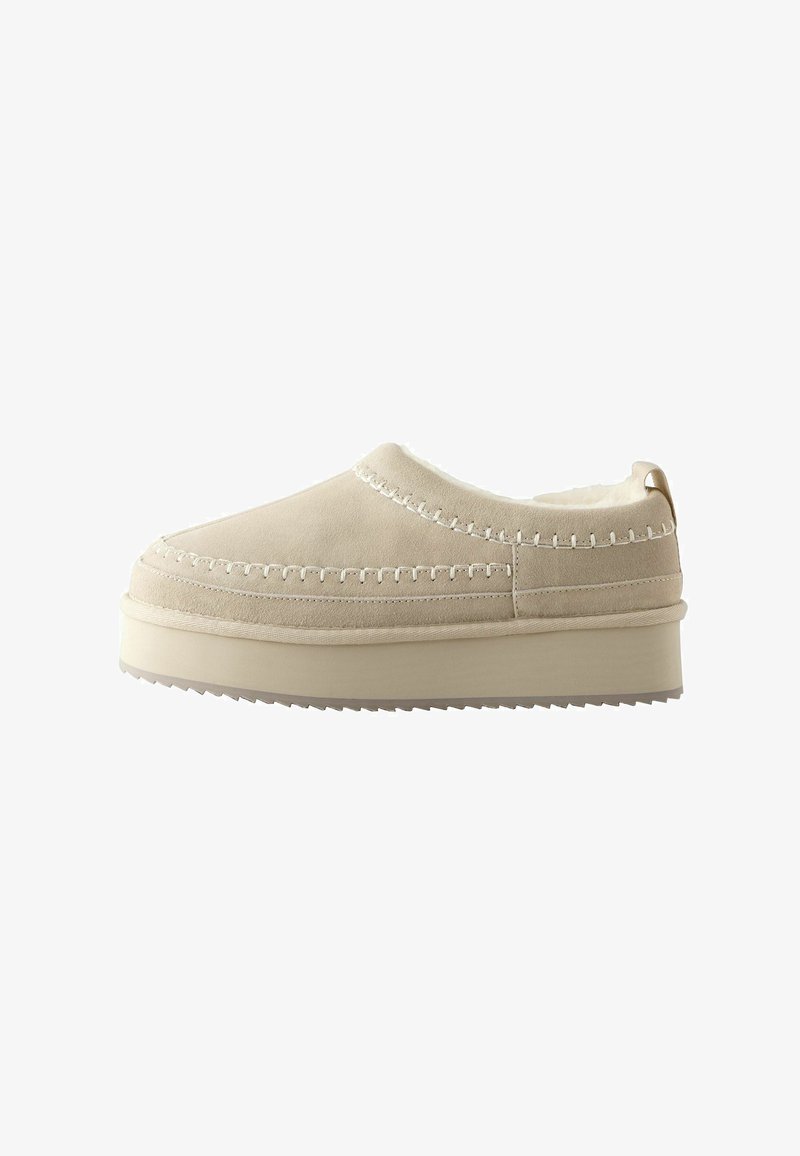 Beige slip-on shoes featuring a soft suede upper, white stitching, and a thick rubber sole with a textured bottom for grip.