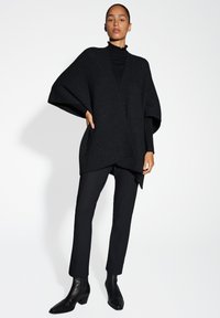 Zwarte gebreide poncho met wijde mouwen, textuurafwerking, gedragen over een zwarte coltrui en gecombineerd met op maat gemaakte zwarte broek en enkellaarsjes.