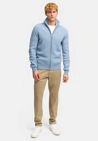 Pull à fermeture éclair bleu clair côtelé avec col montant, associé à un pantalon beige et des baskets blanches. Le design met en valeur un tissu texturé.