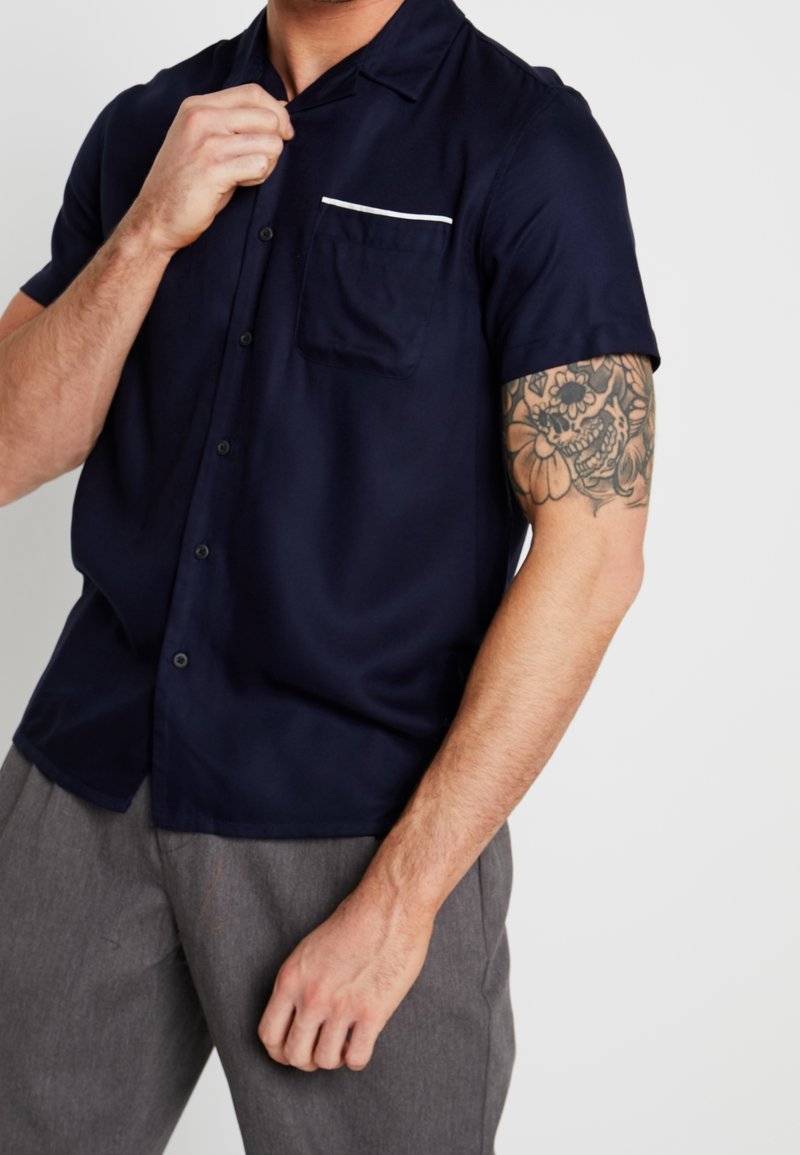 Homme ajustant le col d'une chemise bleu marine à manches courtes boutonnée, montrant un tatouage de crâne floral sur le haut du bras, portant un pantalon gris.
