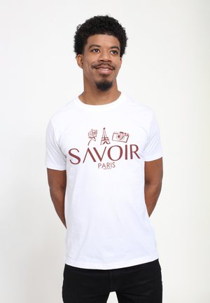 Hombre con camiseta blanca con iconos de rosa, Torre Eiffel y cámara sobre el texto "SAVOIR PARIS", manos detrás de la espalda, sonriendo, fondo claro.