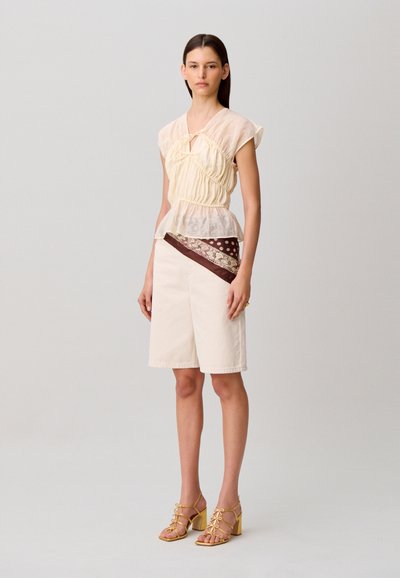 Donna con una maglietta beige trasparente arricciata, pantaloncini bianchi al ginocchio con una fascia marrone a fantasia, e sandali gialli con tacco, in piedi davanti a uno sfondo semplice.