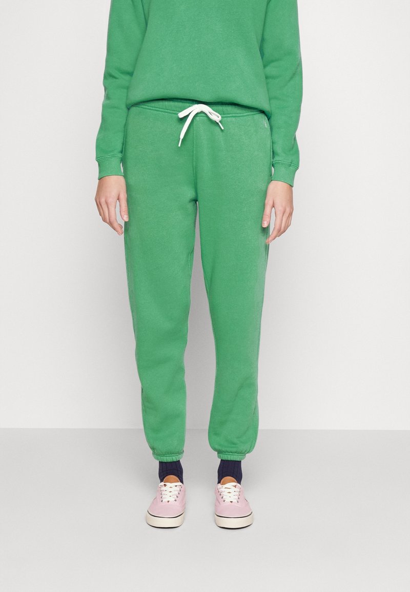 Zelené fleecové jogger kalhoty s elastickým pasem a bílou šňůrkou. Mají zúžené nohavice a boční kapsy. Dohromady s světle růžovými teniskami.