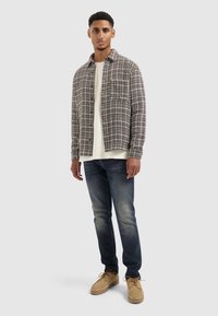 Geruite overhemd in zwart-wit boven een crèmekleurig sweatshirt, gecombineerd met donkere jeans en beige suède schoenen met veters en dikke zolen.