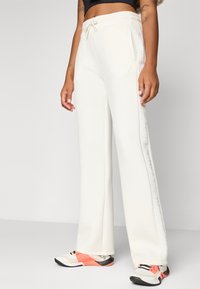 Een persoon staat in witte sweatpants met een zijlogo en kleurrijke sneakers, wat een casual, sportieve outfit laat zien. De achtergrond is neutraal.
