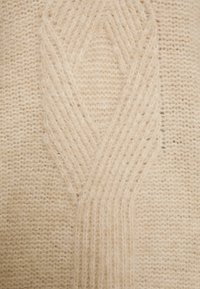 Dzianina w miękkim beżu charakteryzuje się teksturowanym wzorem tworzącym splątane kształty, podkreślającym misterny rzemiosło i warstwowość.