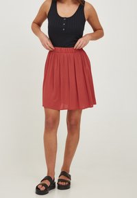 Svart ribbad tanktop med tre knappar, kombinerad med en rostfärgad, knälång plisserad kjol. Bärs med svarta platta sandaler.