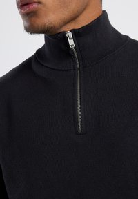 Black knit sweater with a high collar, featuring a half zipper. Texture appears soft and smooth with a consistent, even pattern.
