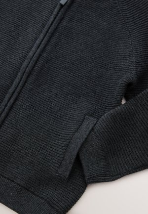 Schwarzer Strickpullover mit Reißverschluss, gerippter Struktur und einer Seitentasche. Der Stoff wirkt dick und weich mit einem strukturierten Design.