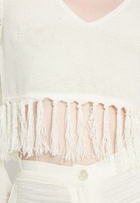 Weißes, gestricktes Crop-Top mit V-Ausschnitt und Fransenverzierung am Saum. Die Textur ist offen gewebt, was ein leichtes, luftiges Erscheinungsbild schafft.