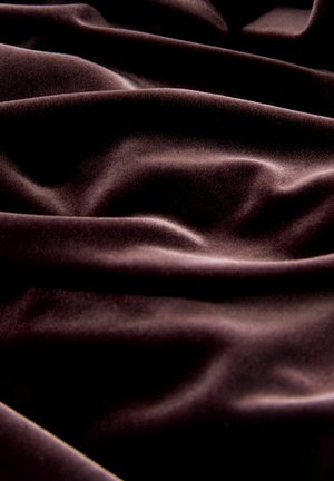 Tissu en velours brun foncé à texture lisse, présentant de profondes plis et rides qui mettent en valeur son drapé luxueux et sa douceur.