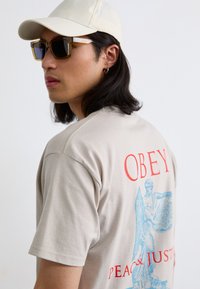 Beige pamut póló, kék grafikai tervezéssel egy angyallal és piros "OBEY PEACE & JUSTICE" felirattal a hátsó részén. A modellen bézs sapka és napszemüveg is található.