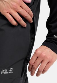Zwarte functionele broek met een ritszak en gestructureerde stof. Het merkembleem, "Jack Wolfskin," is zichtbaar op het been.