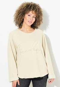 Top de manga larga crema con escote redondeado, presenta "Libertad" en bordado metálico, textura suave y corte relajado. Combinado con jeans oscuros.