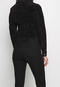 Donna che indossa una giacca nera in suede aderente e jeans neri skinny, mostrata di spalle su uno sfondo bianco semplice.